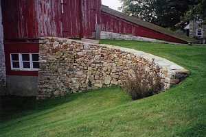 a curved natural stone wall that leads a grass path to an old red barn