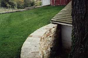 a curved natural stone wall on a grassy hill leading to an old barn