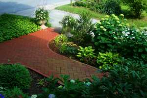 a brick walkway running parallel to a stone driveway and between two gardens full of shrubs