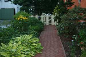 a brick walkway between two gardens leading to a white picket fence