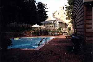 a brick patio around a pool area. a grill is in the foreground with patio chairs and a table in the background