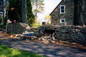 a stone wall that is falling apart with a wheel barrow in front a stone house behind