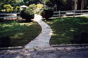 a twisted stone walkway running from a driveway to the backyard between two evergreens and a white fence