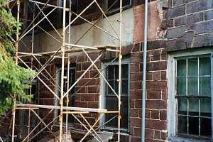 a shirtless man standing on scaffolding next to home with a damaged stone wall